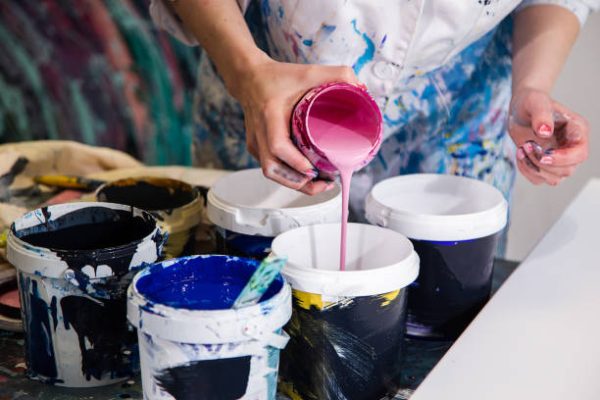 Close up of painter hands pouring bright pink paint from jar into can standing on table next to other cans with paint wearing white coat covered with splashes of paint
